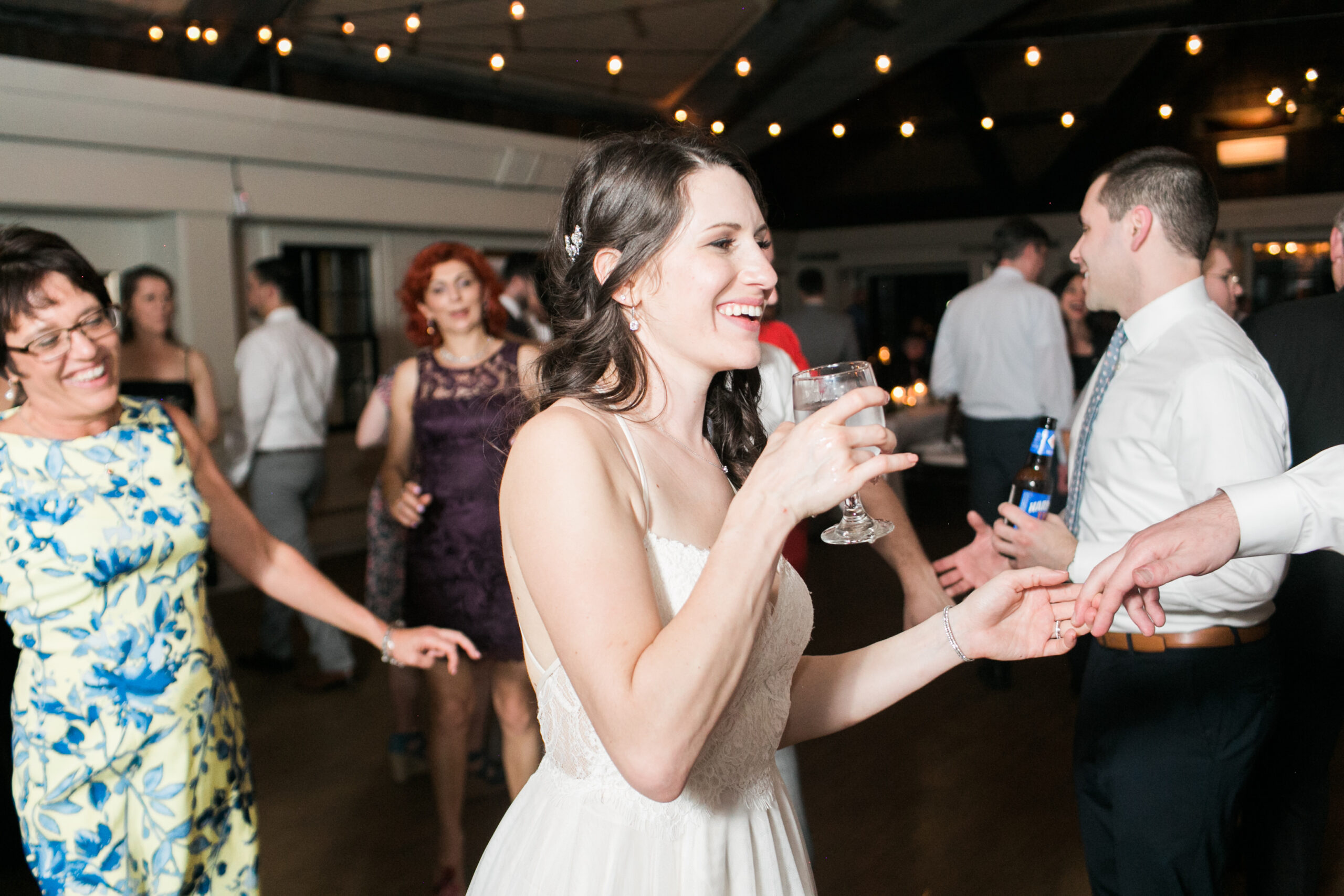 Smiling bride in white dress holding drink on dancefloor with older woman dancing in background with blue and yellow floral dress at Latitude 41 Wedding Mystic with bar lights hung up in background