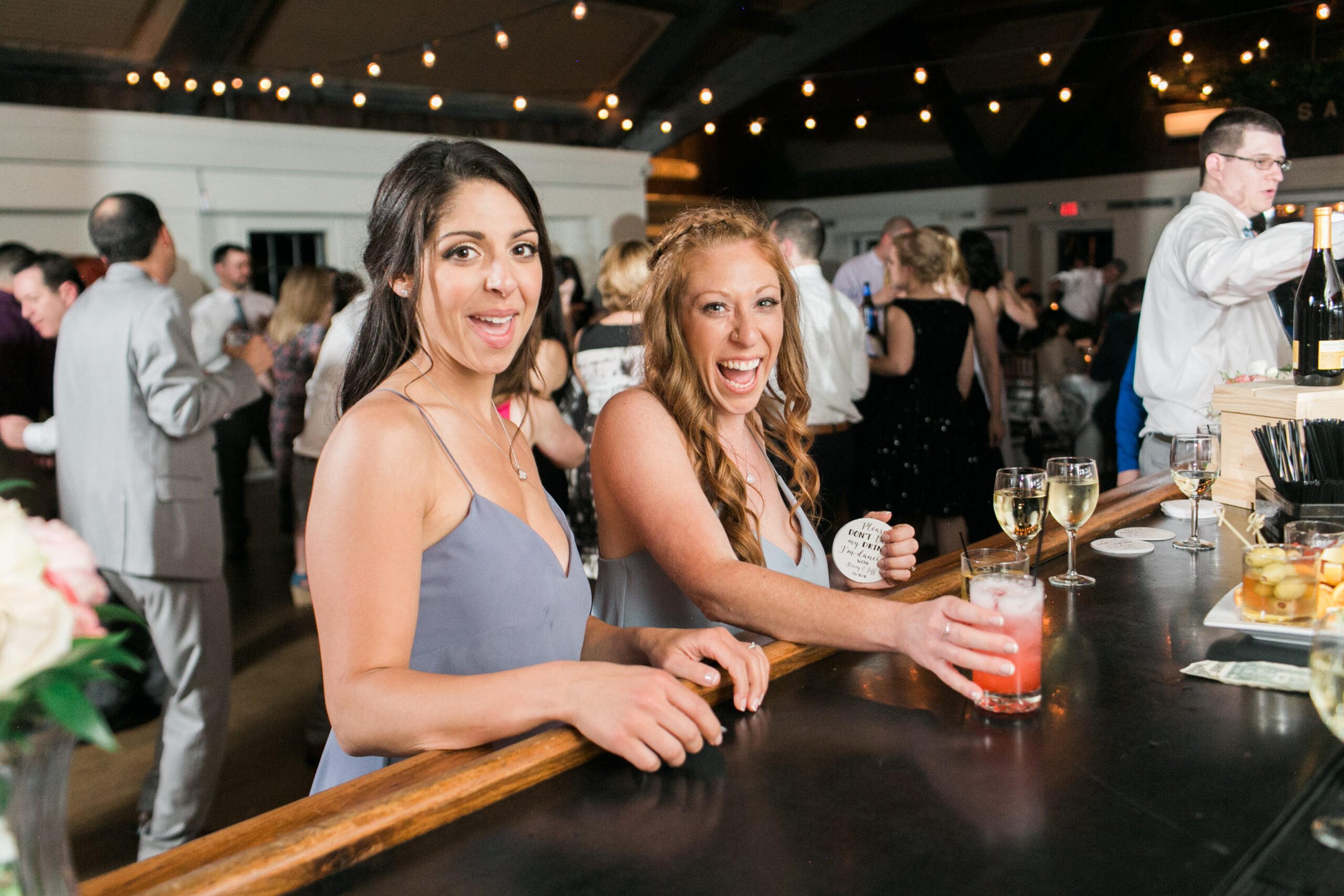 Fun smiling bridesmaids in light blue dresses getting drinks at the bar Latitude 41 Mystic with bar lights hung up in background