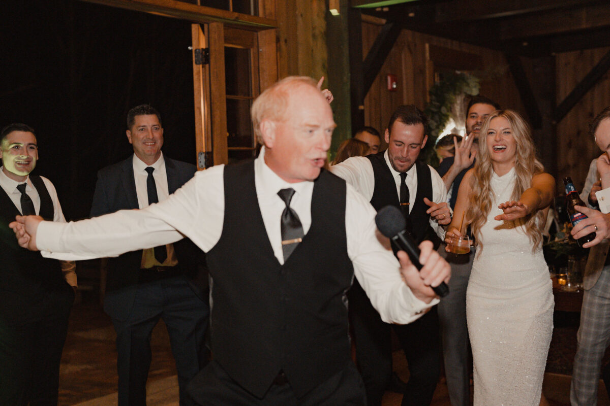 Father of bride holding microphone during lip sync performance at daughters who is in the background at her wedding at The Preserve at Chocorua in Tamworth NH